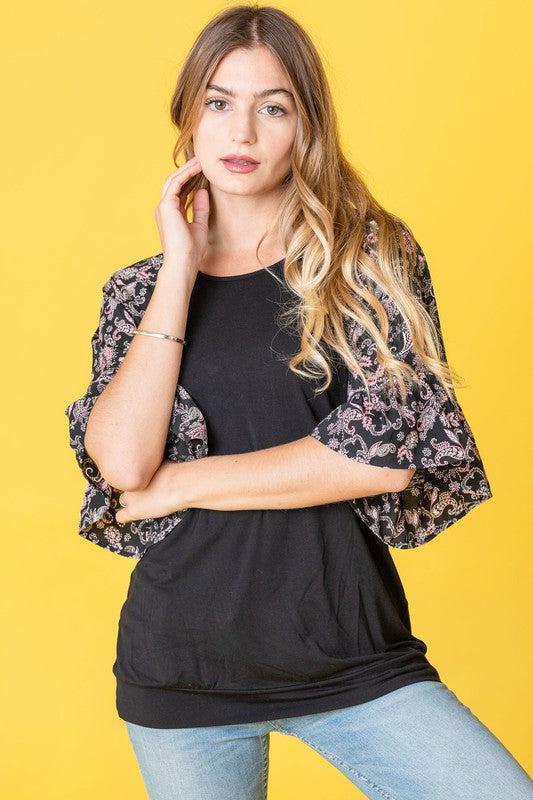 A woman stands against a vibrant yellow background wearing the Ruffle Print Sleeve Tunic, a chic black outfit combining elegance and playful charm. Made in the United States, it features stylish ruffle print sleeves.