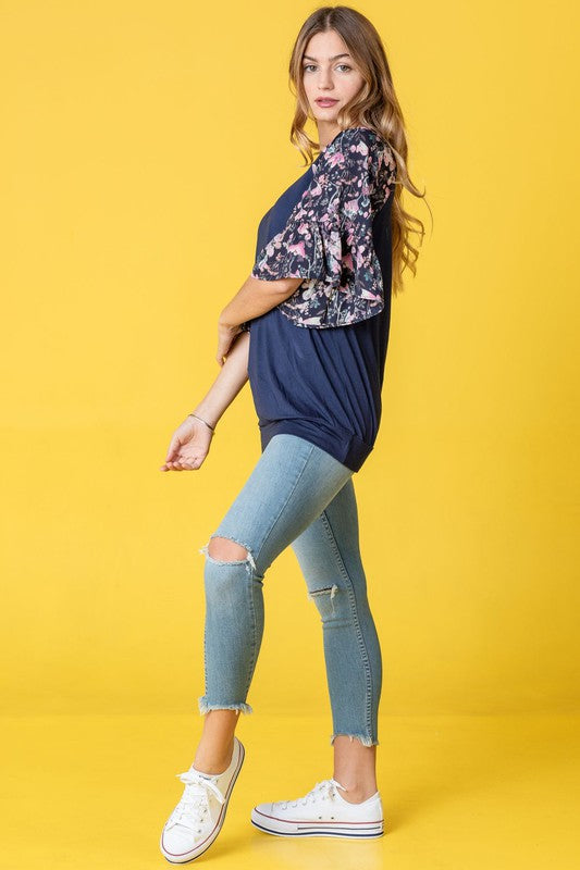 A woman stands against a vibrant yellow background wearing the Ruffle Print Sleeve Tunic, a chic black outfit combining elegance and playful charm. Made in the United States, it features stylish ruffle print sleeves.