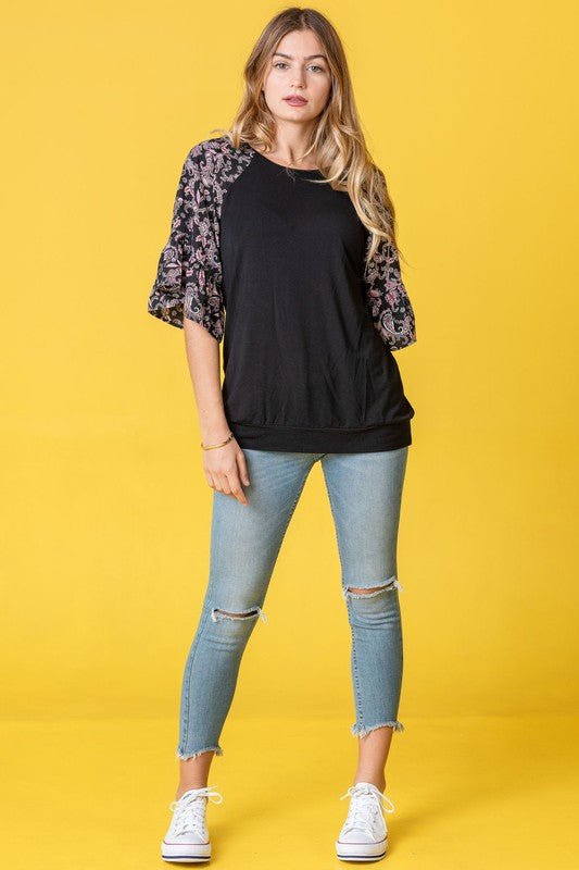 A woman stands against a vibrant yellow background wearing the Ruffle Print Sleeve Tunic, a chic black outfit combining elegance and playful charm. Made in the United States, it features stylish ruffle print sleeves.