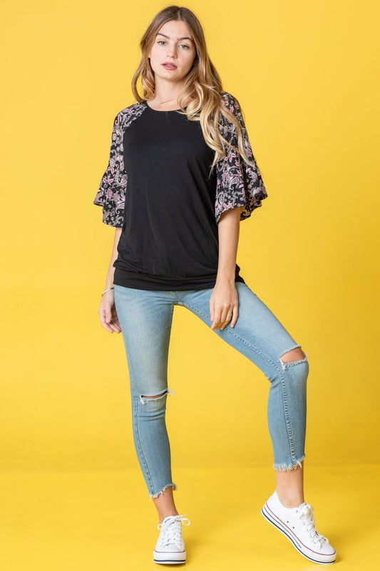 A woman stands against a vibrant yellow background wearing the Ruffle Print Sleeve Tunic, a chic black outfit combining elegance and playful charm. Made in the United States, it features stylish ruffle print sleeves.