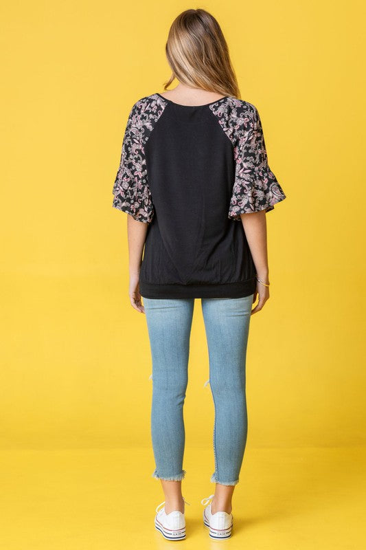 A woman stands against a vibrant yellow background wearing the Ruffle Print Sleeve Tunic, a chic black outfit combining elegance and playful charm. Made in the United States, it features stylish ruffle print sleeves.