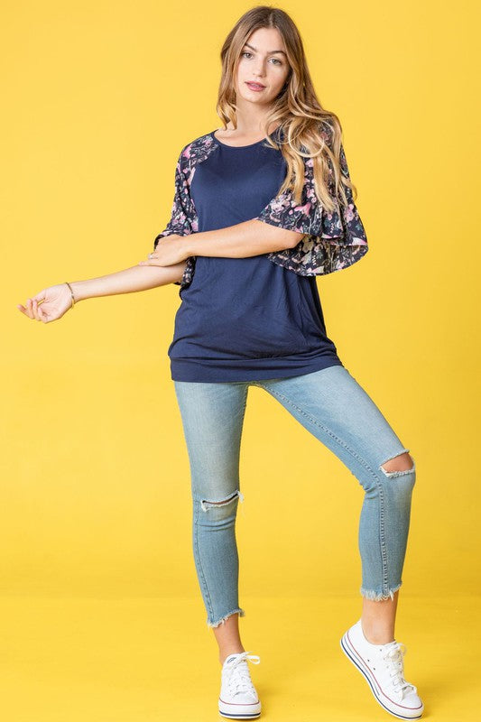 A woman with long hair is wearing the Ruffle Print Sleeve Tunic in navy blue, standing against a yellow background.