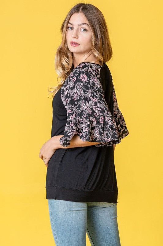 A woman stands against a vibrant yellow background wearing the Ruffle Print Sleeve Tunic, a chic black outfit combining elegance and playful charm. Made in the United States, it features stylish ruffle print sleeves.