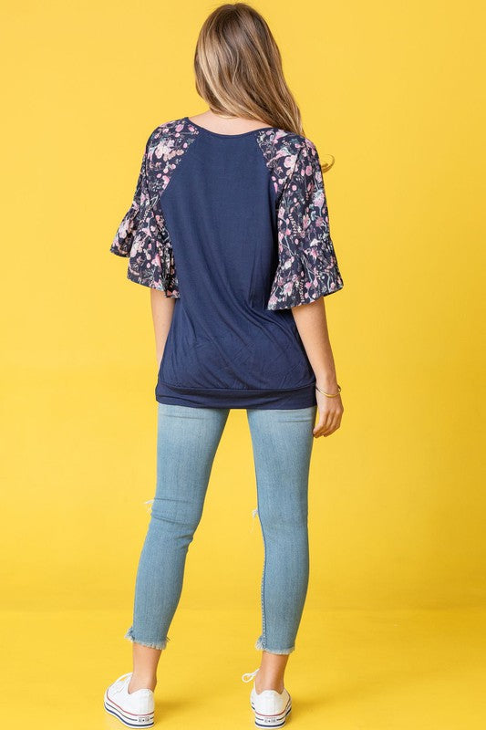 A woman stands against a vibrant yellow background wearing the Ruffle Print Sleeve Tunic, a chic black outfit combining elegance and playful charm. Made in the United States, it features stylish ruffle print sleeves.