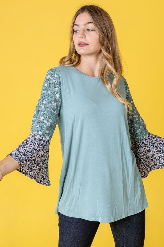 A woman in a Color Block Ruffle Sleeve Tunic, featuring elegant floral bell sleeves, stands against a pink backdrop, gracefully touching her hair.