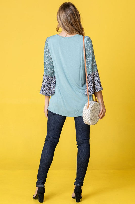 A woman in a Color Block Ruffle Sleeve Tunic, featuring elegant floral bell sleeves, stands against a pink backdrop, gracefully touching her hair.