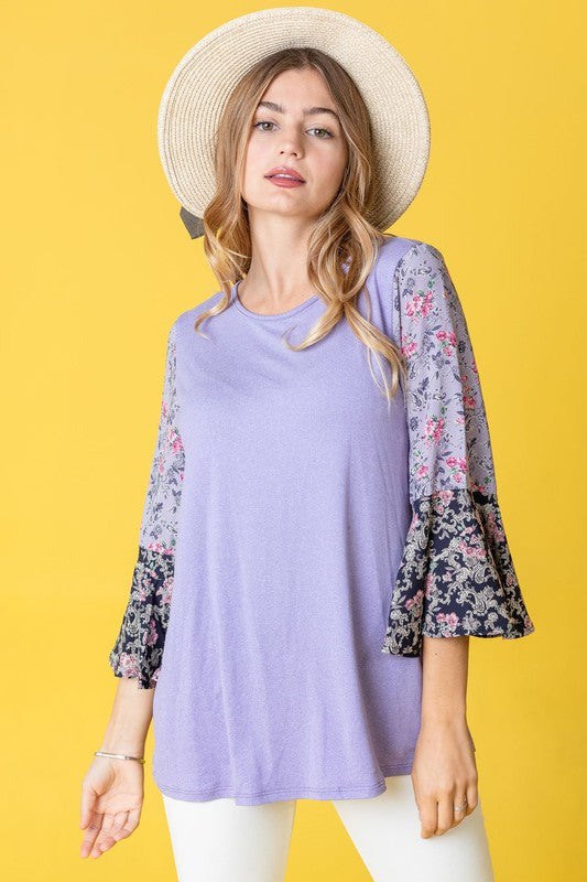 A woman in a Color Block Ruffle Sleeve Tunic, featuring elegant floral bell sleeves, stands against a pink backdrop, gracefully touching her hair.