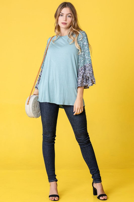 A woman in a Color Block Ruffle Sleeve Tunic, featuring elegant floral bell sleeves, stands against a pink backdrop, gracefully touching her hair.