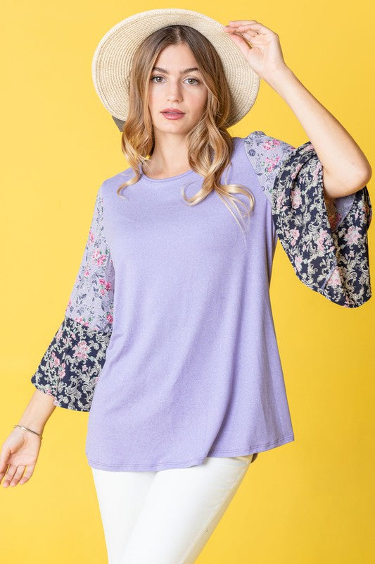 A woman in a Color Block Ruffle Sleeve Tunic, featuring elegant floral bell sleeves, stands against a pink backdrop, gracefully touching her hair.