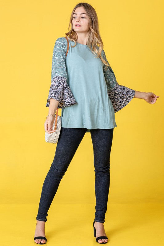 A woman in a Color Block Ruffle Sleeve Tunic, featuring elegant floral bell sleeves, stands against a pink backdrop, gracefully touching her hair.