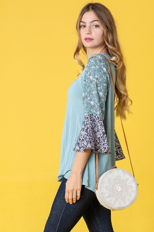 A woman in a Color Block Ruffle Sleeve Tunic, featuring elegant floral bell sleeves, stands against a pink backdrop, gracefully touching her hair.