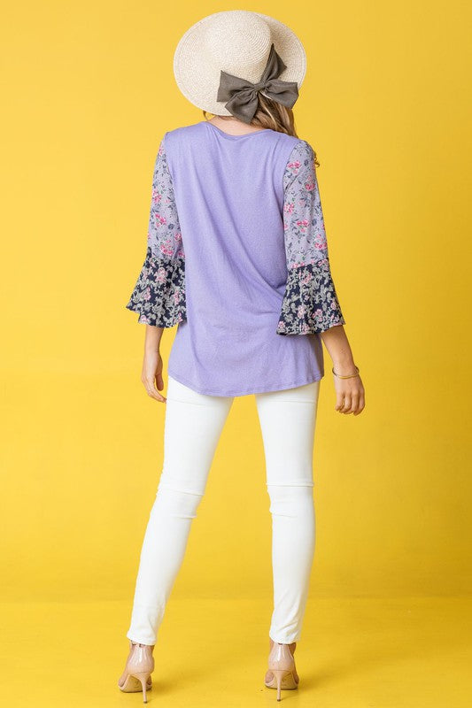 A woman in a Color Block Ruffle Sleeve Tunic, featuring elegant floral bell sleeves, stands against a pink backdrop, gracefully touching her hair.