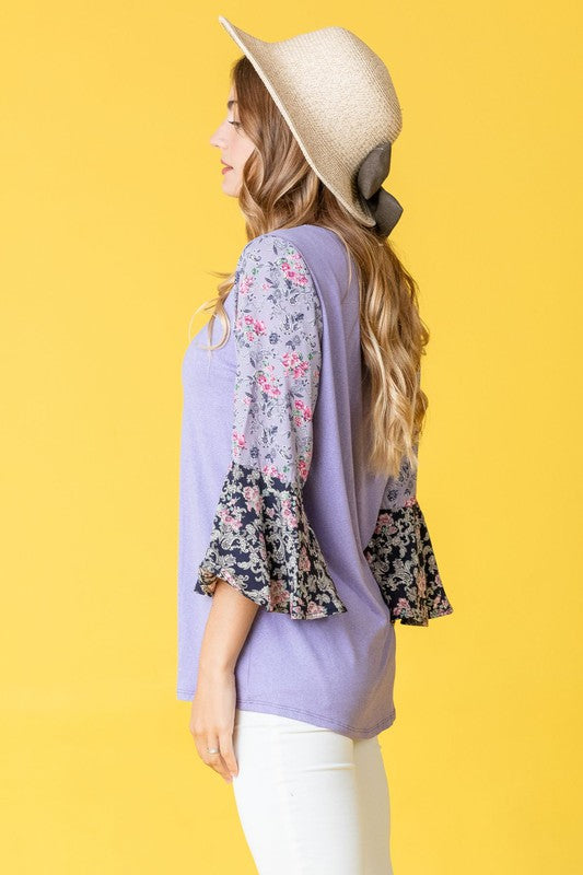 A woman in a Color Block Ruffle Sleeve Tunic, featuring elegant floral bell sleeves, stands against a pink backdrop, gracefully touching her hair.