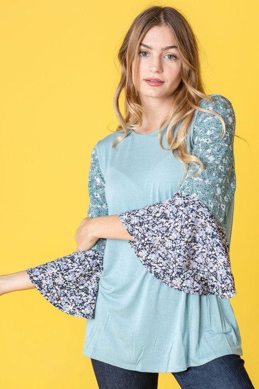 A woman in a Color Block Ruffle Sleeve Tunic, featuring elegant floral bell sleeves, stands against a pink backdrop, gracefully touching her hair.