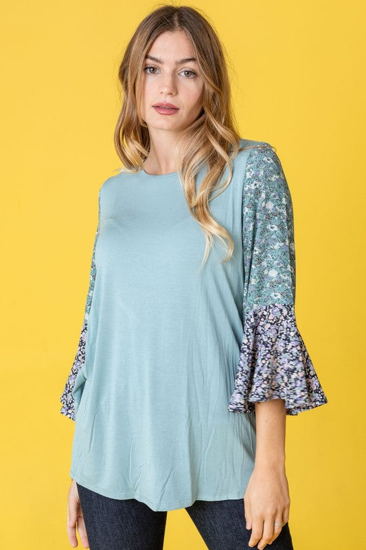 A woman in a Color Block Ruffle Sleeve Tunic, featuring elegant floral bell sleeves, stands against a pink backdrop, gracefully touching her hair.