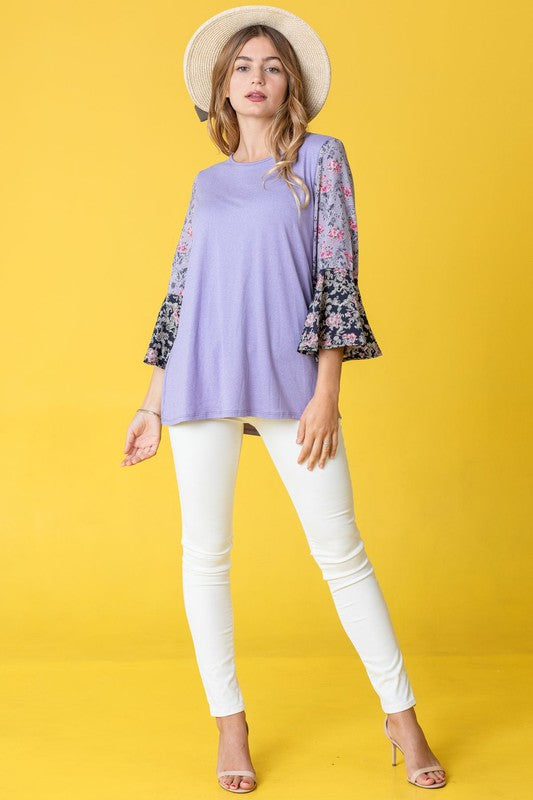 A woman in a Color Block Ruffle Sleeve Tunic, featuring elegant floral bell sleeves, stands against a pink backdrop, gracefully touching her hair.