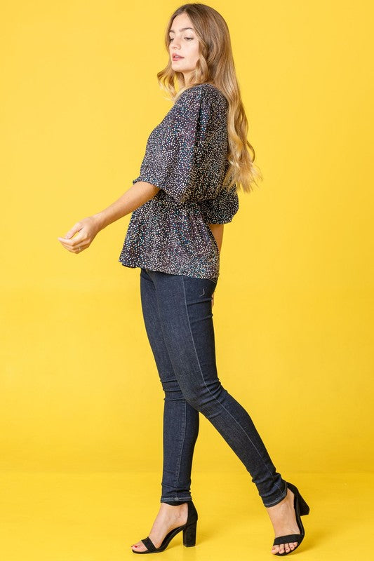 A woman with long dark hair is wearing a Plus Polka Dot Square Neck Peplum Top and blue jeans, standing in front of a beige curtain.