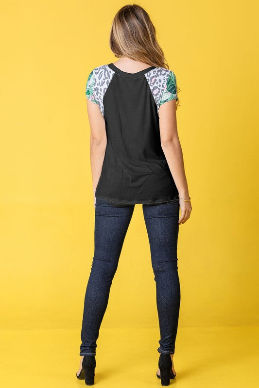 A woman in a Leopard Tropical Cap Sleeve Color Block Tunic and dark jeans stands against a vibrant yellow background.