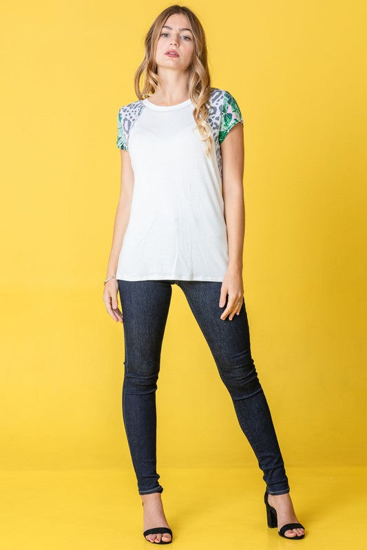 A woman in a Leopard Tropical Cap Sleeve Color Block Tunic and dark jeans stands against a vibrant yellow background.