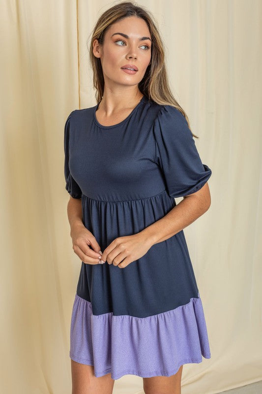 A woman wearing the Two Tone Tiered Mini Dress, featuring a navy top and lavender bottom, stands against a neutral backdrop. She pairs it with black strapped shoes.