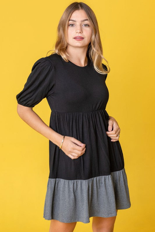 A woman wearing the Two Tone Tiered Mini Dress, featuring a navy top and lavender bottom, stands against a neutral backdrop. She pairs it with black strapped shoes.