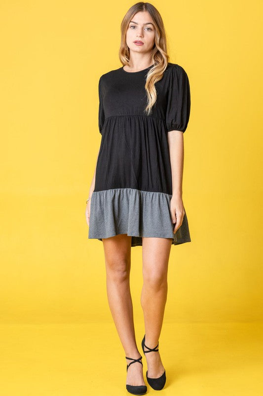 A woman wearing the Two Tone Tiered Mini Dress, featuring a navy top and lavender bottom, stands against a neutral backdrop. She pairs it with black strapped shoes.