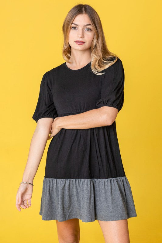 A woman wearing the Two Tone Tiered Mini Dress, featuring a navy top and lavender bottom, stands against a neutral backdrop. She pairs it with black strapped shoes.