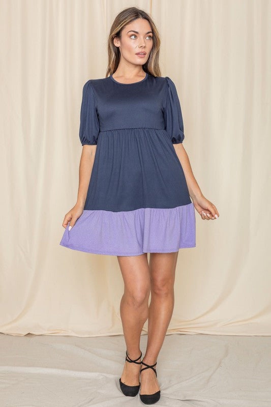 A woman wearing the Two Tone Tiered Mini Dress, featuring a navy top and lavender bottom, stands against a neutral backdrop. She pairs it with black strapped shoes.