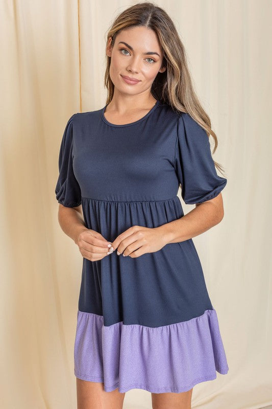 A woman wearing the Two Tone Tiered Mini Dress, featuring a navy top and lavender bottom, stands against a neutral backdrop. She pairs it with black strapped shoes.