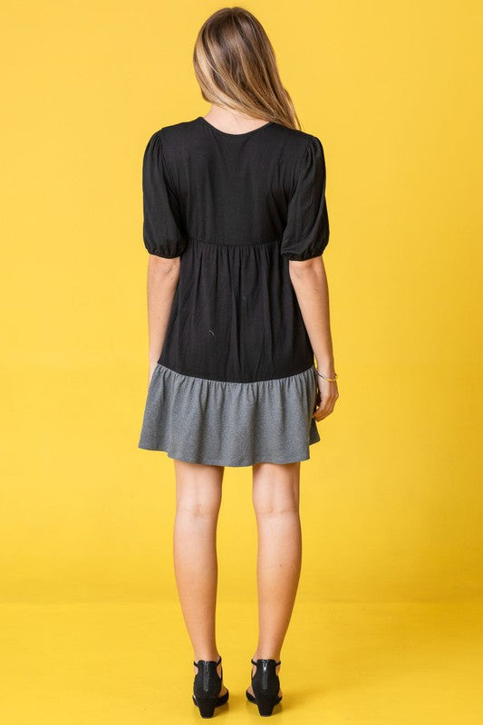 A woman wearing the Two Tone Tiered Mini Dress, featuring a navy top and lavender bottom, stands against a neutral backdrop. She pairs it with black strapped shoes.