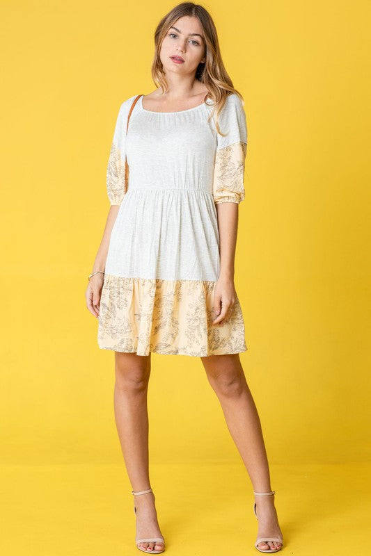 A person stands against a yellow background, wearing the Square Neck Floral Mini Dress, which features patterns on the sleeves and hem. The square neckline adds a touch of elegance, perfectly capturing the breezy fashion essence popular in the United States.