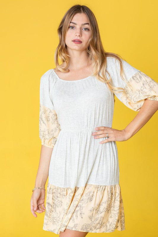 A person stands against a yellow background, wearing the Square Neck Floral Mini Dress, which features patterns on the sleeves and hem. The square neckline adds a touch of elegance, perfectly capturing the breezy fashion essence popular in the United States.