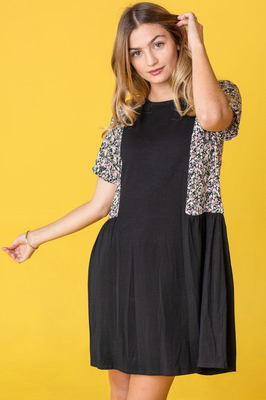 A person stands against a yellow background, wearing the Ditsy Floral Contrast Mini Dress, featuring navy short sleeves and side panels with a ditsy floral pattern.