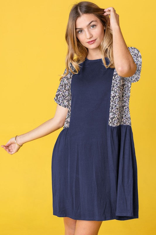 A person stands against a yellow background, wearing the Ditsy Floral Contrast Mini Dress, featuring navy short sleeves and side panels with a ditsy floral pattern.