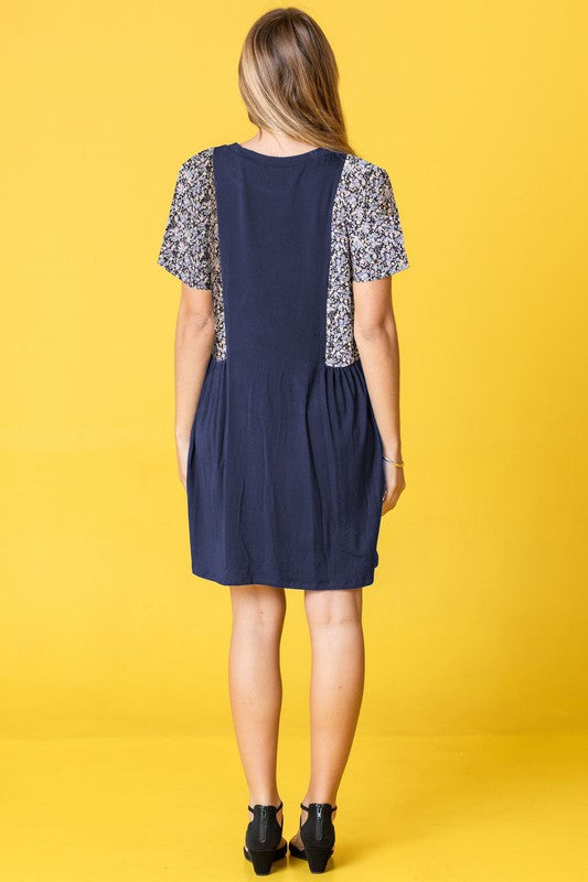 A person stands against a yellow background, wearing the Ditsy Floral Contrast Mini Dress, featuring navy short sleeves and side panels with a ditsy floral pattern.