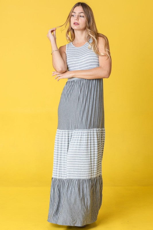 A woman confidently stands against a vibrant yellow background, wearing the Sleeveless Stripe Tiered Maxi Dress, made in the United States.