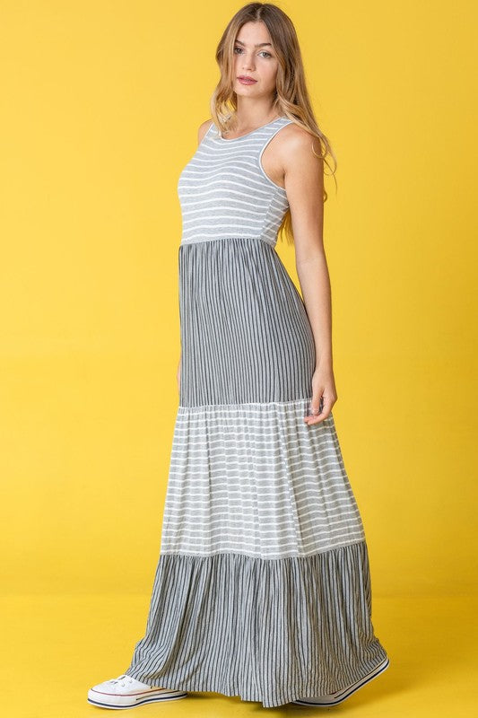 A person stands against a pink backdrop wearing the Plus Sleeveless Stripe Tiered Maxi Dress, made from a comfortable rayon spandex blend with alternating white and navy blue stripes, paired with white sneakers.