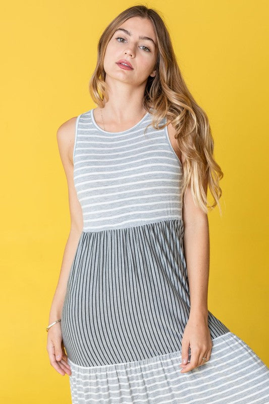 A woman confidently stands against a vibrant yellow background, wearing the Sleeveless Stripe Tiered Maxi Dress, made in the United States.