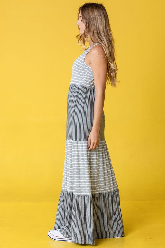 A person stands against a pink backdrop wearing the Plus Sleeveless Stripe Tiered Maxi Dress, made from a comfortable rayon spandex blend with alternating white and navy blue stripes, paired with white sneakers.