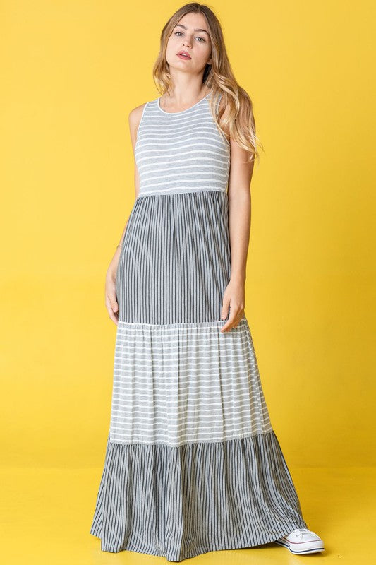 A person stands against a pink backdrop wearing the Plus Sleeveless Stripe Tiered Maxi Dress, made from a comfortable rayon spandex blend with alternating white and navy blue stripes, paired with white sneakers.