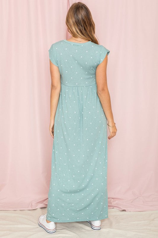 A woman in a chic Polka Dot Folded Cap Sleeve Maxi Dress stands gracefully against a pink curtain backdrop, exuding effortless elegance reminiscent of timeless United States style.