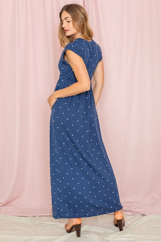 A woman in a chic Polka Dot Folded Cap Sleeve Maxi Dress stands gracefully against a pink curtain backdrop, exuding effortless elegance reminiscent of timeless United States style.