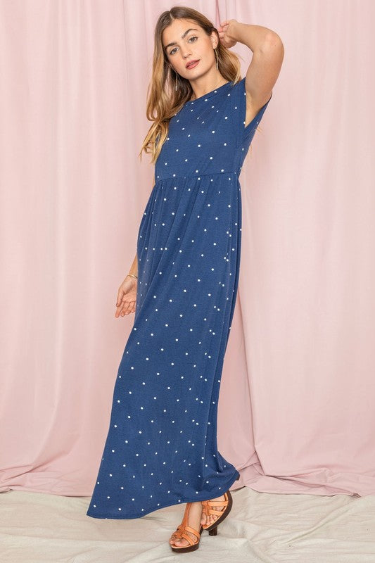 A woman in a chic Polka Dot Folded Cap Sleeve Maxi Dress stands gracefully against a pink curtain backdrop, exuding effortless elegance reminiscent of timeless United States style.