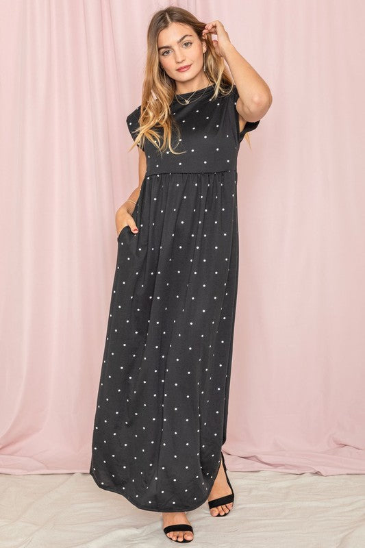 A woman in a chic Polka Dot Folded Cap Sleeve Maxi Dress stands gracefully against a pink curtain backdrop, exuding effortless elegance reminiscent of timeless United States style.