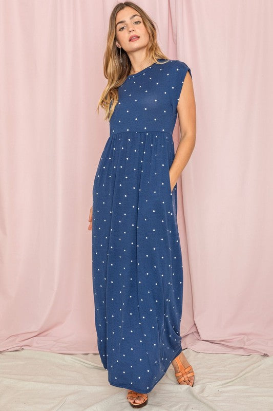 A woman in a chic Polka Dot Folded Cap Sleeve Maxi Dress stands gracefully against a pink curtain backdrop, exuding effortless elegance reminiscent of timeless United States style.