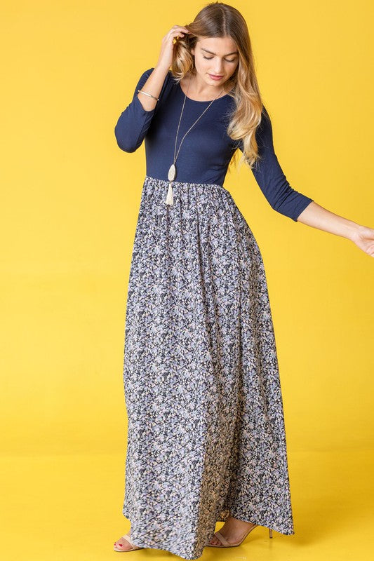 A woman in a blue top and the Ditsy Floral Maxi Dress stands against a yellow background, wearing a necklace and holding her hair.