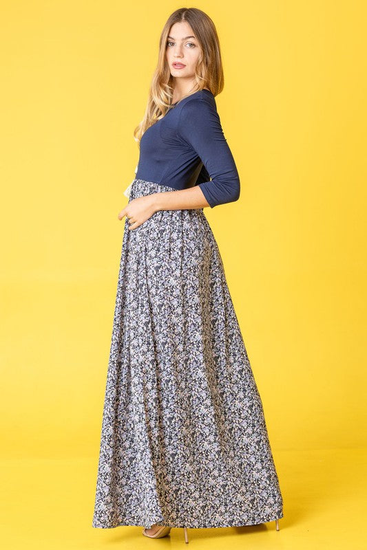 A woman elegantly stands in a room with wooden flooring and modern furniture, wearing a ditsy floral maxi dress and a long-sleeve navy top.