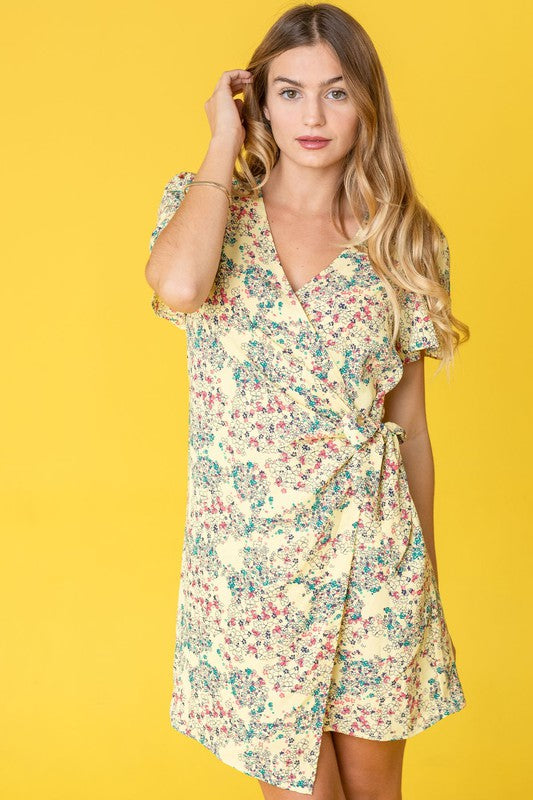 Elegantly perched on a round chair, the woman wears the Ditsy Floral Wrap Dress in a vibrant yellow, featuring a charming design crafted in the United States.