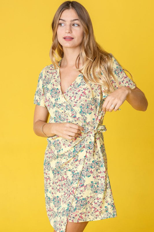 Elegantly perched on a round chair, the woman wears the Ditsy Floral Wrap Dress in a vibrant yellow, featuring a charming design crafted in the United States.