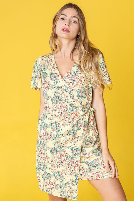 Wearing the vibrant Ditsy Floral Wrap Dress made from polyester, a person stands gracefully against a cheerful yellow background. Crafted in the United States, this eye-catching ensemble showcases both elegance and quality craftsmanship.